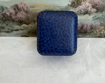 May include: A small, square-shaped jewelry box featuring a textured, deep blue exterior with a mottled, pebbled finish. The box has rounded corners and a smooth, uniform appearance, designed to hold small items securely within its compact frame.