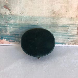 May include: A dark green, round velvet jewelry box. The box is closed and has a small, gold-colored clasp. The background is a faded, textured painting with shades of blue, green, and white.