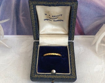 Puede incluir: Un anillo de oro fino con un patrón grabado dentro de un joyero forrado de terciopelo azul oscuro. La tapa interior presenta el texto impreso W. F. ARNETT Jeweler Longview, Washington, enmarcado por un borde con diseño de greca azul y dorada.