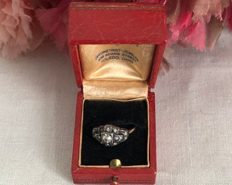 Puede incluir: Un anillo de tono plateado con varias piedras claras pequeñas engastadas en una cabeza rectangular y geométrica, dentro de un joyero rojo rectangular. La tapa interior presenta el texto: OPTOMETRIST-JEWELER 508 ADAMS STREET TOLEDO, OHIO.