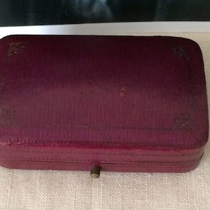 May include: A rectangular, burgundy-colored jewelry box with a textured surface. The box has a small, brass-colored clasp and decorative corner accents. The box is closed and sits on a light-colored, textured surface.