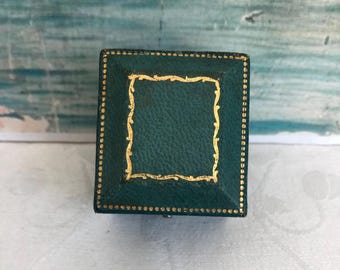 May include: A square, dark green jewelry box with a textured surface. The lid features a decorative gold border along the edges and a central rectangular frame with a wavy, ornate gold pattern embossed on the top surface of the box.