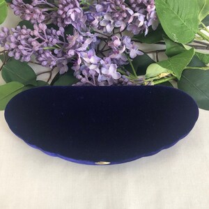 May include: A deep blue velvet eyeglass case with a gold-colored clasp. The case has a rounded shape and is set against a backdrop of purple flowers and green leaves.