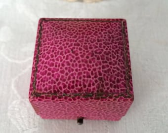 Puede incluir: Un joyero cuadrado con un exterior texturizado de color magenta que presenta un patrón granulado distintivo. La tapa tiene una parte superior ligeramente abovedada con bordes cosidos oscuros y un pequeño cierre metálico en la parte frontal.