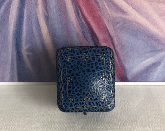 Puede incluir: Un joyero rectangular con un exterior azul texturizado que presenta un patrón moteado y delicados detalles de filigrana de color dorado a lo largo de los bordes. La tapa tiene bisagras y muestra un diseño de borde rectangular central en tinta dorada.