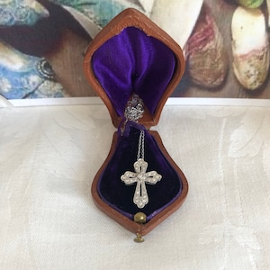 May include: A silver cross pendant necklace displayed in a vintage brown leather jewelry box with a purple velvet interior. The cross is adorned with small, sparkling stones. The box is open, showcasing the necklace against the plush background.