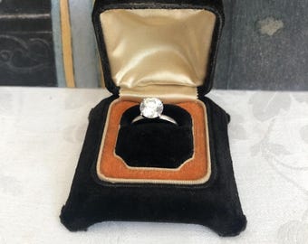 May include: A silver-toned ring featuring a single round, clear faceted stone set in a prong setting. The ring is displayed inside a black velvet-lined jewelry box with a cream-colored satin interior lid and an orange velvet base insert.