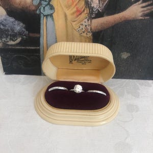 May include: An open, off-white jewelry box with a maroon velvet interior holds three rings. The center ring features a large, round, clear stone. The box is labeled "Jewelers and Optometrists, Fond du Lac, Wis."