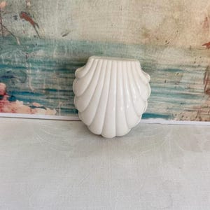 May include: A glossy white ceramic or plastic object shaped like a scallop shell. The item features a series of vertical, radiating ridges that extend from the base to the rounded top edge, creating a symmetrical, fan-like appearance.