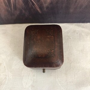 May include: A rectangular dark brown jewelry box with a textured leather-like finish and a decorative embossed border pattern on the lid. The box features a small metal latch on the front edge and has a rounded, slightly domed top surface.
