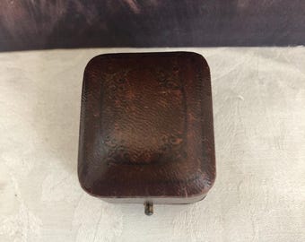 Puede incluir: Un joyero rectangular de color marrón oscuro con un acabado texturizado similar al cuero y un patrón de borde en relieve decorativo en la tapa. La caja presenta un pequeño cierre de metal en el borde frontal y una tapa ligeramente abombada.