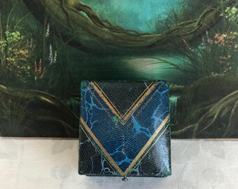 May include: A square jewelry box featuring a textured dark blue and black marbled finish with a prominent gold-colored V-shaped geometric pattern on the lid. The box has a small metal latch at the bottom center and a structured, rigid exterior.