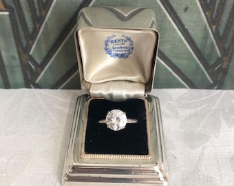 May include: A round-cut solitaire diamond ring with a silver-toned band, presented in a rectangular silver-toned jewelry box with a black velvet insert. The interior lid features a blue logo reading Kents Limited Jewelers Toronto.