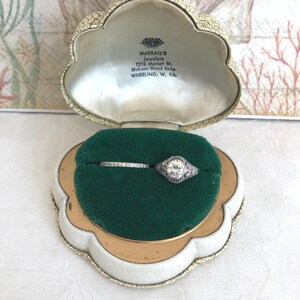 May include: A silver-toned engagement ring with a central round stone and a matching thin band, both set in a shell-shaped jewelry box. The interior lid features the text: McGRAIL'S Jewelers, 1212 Market St., McLure Hotel Bldg., WHEELING, W. VA.