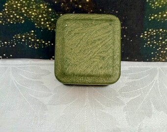 Puede incluir: Un recipiente metálico de forma cuadrada con un acabado verde oliva texturizado y brillante. La tapa presenta un sutil patrón de remolinos en toda su superficie y los bordes son redondeados, lo que le da un aspecto compacto a esta caja.