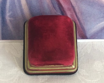 May include: A rectangular jewelry box featuring a deep red velvet top cushion. The base of the box is constructed with tiered edges finished in a muted gold tone, while the bottom perimeter is lined with a thin band of black material.