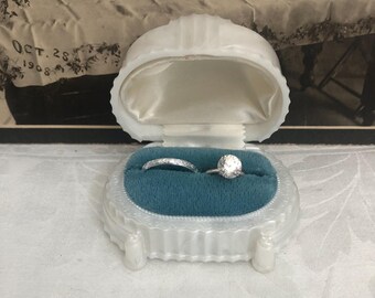 Puede incluir: Un joyero blanco de forma ovalada, estilo concha, con un interior de terciopelo azul verdoso. Dentro de la caja, hay una alianza plateada con un patrón grabado y un anillo plateado con una piedra grande, redonda y transparente.