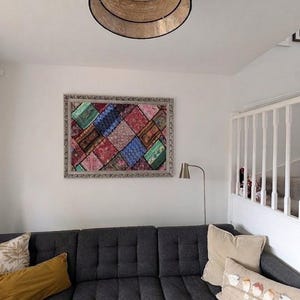 May include: A dark grey tufted sectional sofa with various throw pillows, including a long rectangular one with a bird graphic. Above the sofa hangs a rectangular wall art piece featuring a colourful patchwork pattern in a silver frame.