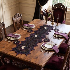 May include: A long, black and purple crocheted table runner featuring a repeating crescent moon motif and intricate web-like patterns. The runner is centered on a wooden dining table, accompanied by matching circular crocheted coasters.