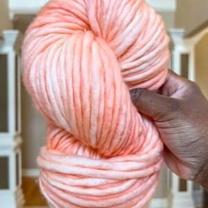 May include: A large skein of super chunky yarn in a soft peach color with white highlights. The yarn is thick and appears soft, ideal for arm knitting or creating large-scale projects. The texture is clearly visible, suggesting a luxurious feel.