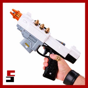 May include: A toy blaster featuring a white and grey body with an orange muzzle, three gold-coloured conical accents on top, and a black textured grip. A yellow warning label with a heat symbol is visible on the side of the grey component.
