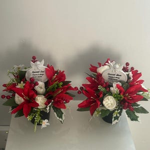 Pode incluir: Dois arranjos florais em vasos pretos com lírios vermelhos, rosas brancas, folhagem verde e bagas vermelhas. Cada arranjo inclui uma placa oval branca com uma figura de anjo, com a inscrição "Special Dad" ou "Special Mum".