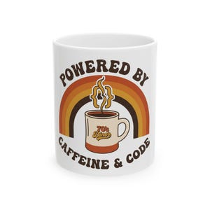 Peut inclure: Un mug en céramique blanche orné d'un graphique représentant une tasse de café avec de la vapeur en forme d'accolades. Au-dessus de la tasse se trouve un arc-en-ciel. Le texte indique "POWERED BY CAFFEINE & CODE" et "70's Diner" sur la tasse.