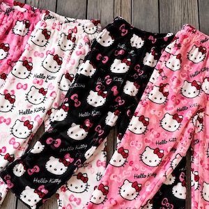 Puede incluir: Cuatro pares de pantalones de estar por casa con un patrón repetitivo de Hello Kitty, lazos y texto. Los pantalones vienen en colores rosa, blanco y negro, todos con cintura elástica. Cada par muestra la cara de Hello Kitty y el texto Hello Kitty impreso sobre la tela suave en un diseño repetitivo constante.