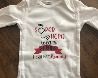 Op de afbeelding: Witte babybody met lange mouwen. De tekst "My Super Hero wears Scrubs, I call her Mommy" is in rood en zwart gedrukt. Een stethoscoopafbeelding is ook inbegrepen. De body is van Gerber en is voor baby's van 6-9 maanden.