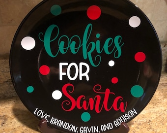 May include: A black ceramic plate with the words "Cookies for Santa" in teal and red script. The plate is decorated with white, red, and teal polka dots and includes the names "Love, Brandon, Gavin, and Addison" in white text.
