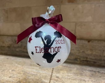 May include: White Christmas bauble with a glitter finish featuring a black silhouette of a cheerleader with pom-poms. The bauble has red and white stars and the text "Eleanor" and "2021".