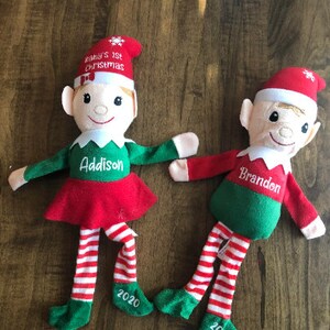 May include: Two personalized Christmas elf dolls with red and green outfits. The girl elf is wearing a red dress and the boy elf is wearing a green shirt. Both elves have white striped stockings and green shoes. The girl elf's dress says "Addison" and the boy elf's shirt says "Brandon". Both elves have a red hat with the text "Baby's 1st Christmas" and the year "2020" on their shoes.