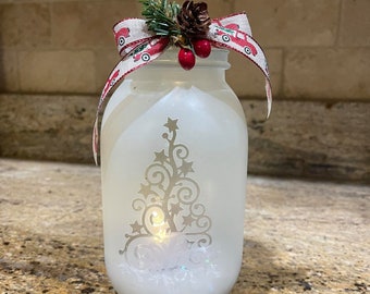 May include: A white frosted glass jar with a silver Christmas tree design and a red and white ribbon with a pine sprig and pine cone on top.
