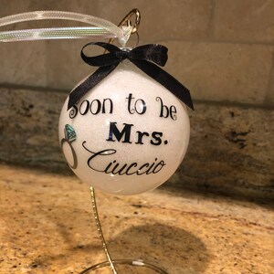 May include: A white glittery Christmas ornament with the text "soon to be Mrs. Cuccio" in black script. A black ribbon and a silver and white ribbon are attached to the top. A diamond ring graphic is also on the ornament.
