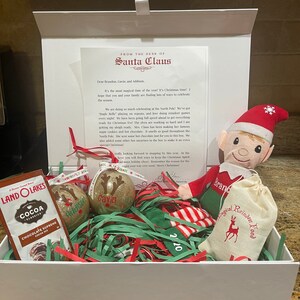 May include: A white gift box filled with Christmas-themed items, including a small elf doll, a bag of reindeer food, two glittery ornaments, and a package of cocoa mix. The box is decorated with red and green tissue paper and a note from Santa Claus.