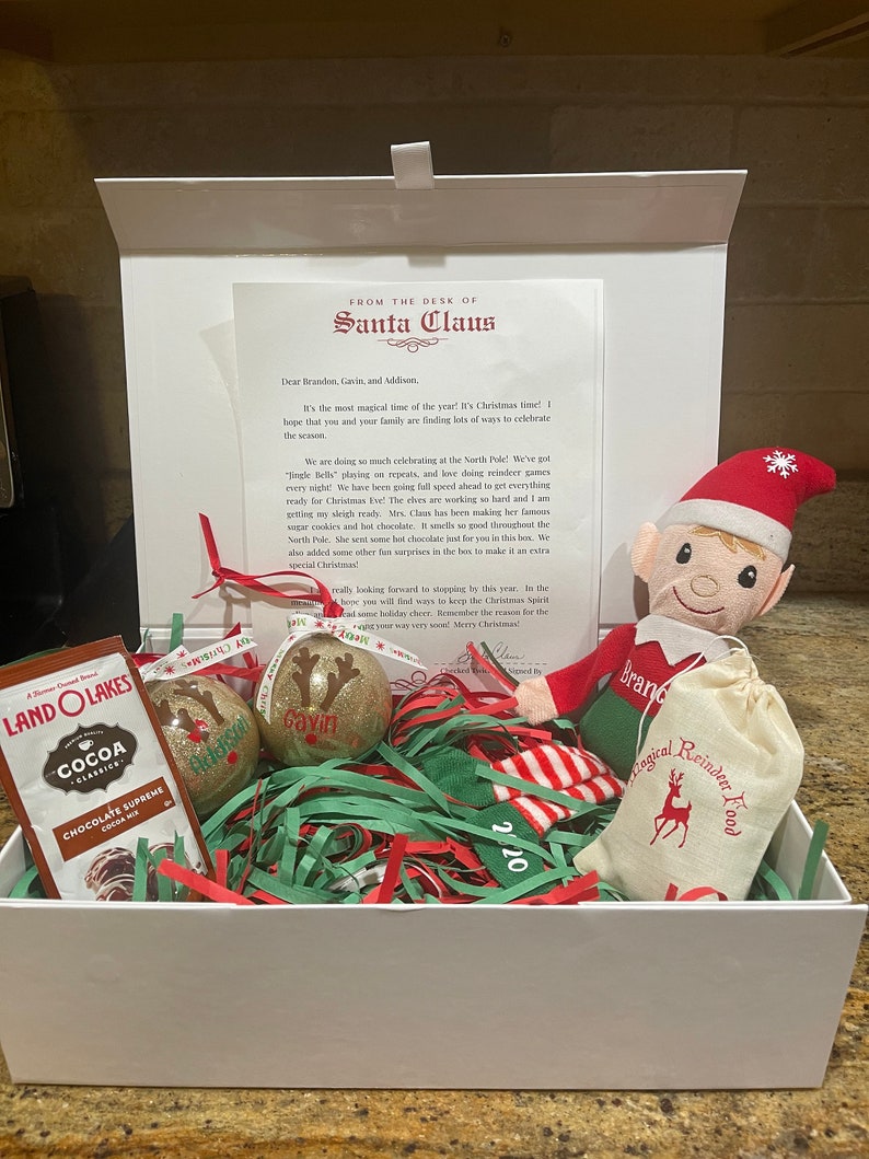 May include: A white gift box filled with Christmas-themed items, including a small elf doll, a bag of reindeer food, two glittery ornaments, and a package of cocoa mix. The box is decorated with red and green tissue paper and a note from Santa Claus.