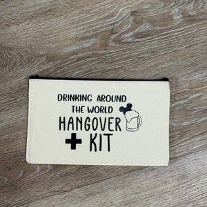 Könnte beinhalten: Beige Canvas-Reißverschlusstasche mit schwarzem Text: "DRINKING AROUND THE WORLD HANGOVER + KIT". Rechts vom Text befindet sich eine Cartoon-Bierkrug-Illustration mit Schaum und Micky-Maus-Ohren.
