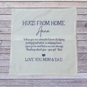 May include: A light green pillowcase with the text "HUGS FROM HOME" and the name "Anna" in dark blue. The pillowcase includes a message from "Mom & Dad" encouraging the recipient. The pillowcase is on a wooden surface.