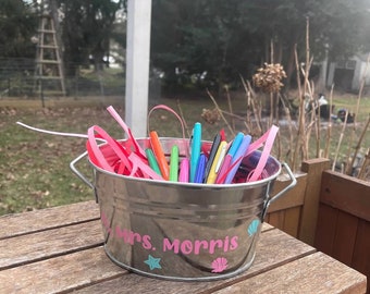 May include: A silver metal bucket filled with pens and pink ribbons. The bucket is personalised with the name "Mrs. Morris" in pink lettering, along with a starfish and seashell design. The bucket sits on a wooden surface.