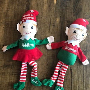 Può includere: Due bambole di elfo di peluche, una in rosso e verde con il nome Addison e l'anno 2020, l'altra in verde e bianco con il nome Brandon e l'anno 2020. Entrambe le bambole hanno un cappello rosso con un fiocco di neve in cima e il testo "Baby's 1st Christmas".