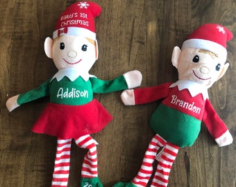 May include: Two plush elf dolls, one in red and green with the name Addison and the year 2020, the other in green and white with the name Brandon and the year 2020. Both dolls have a red hat with a snowflake on top and the text "Baby's 1st Christmas".