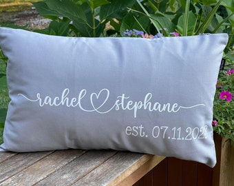 May include: A rectangular gray pillow with white script text that reads "rachel ♡ stephane" above "est. 07.11.2023". The pillow is on a wooden surface, with green foliage and flowers in the background.