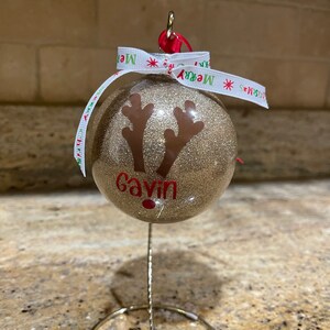 May include: Gold glitter Christmas ornament with a reindeer silhouette and the name "Gavin" written in red. The ornament is on a metal stand with a white ribbon with the words "Merry Christmas" printed on it.