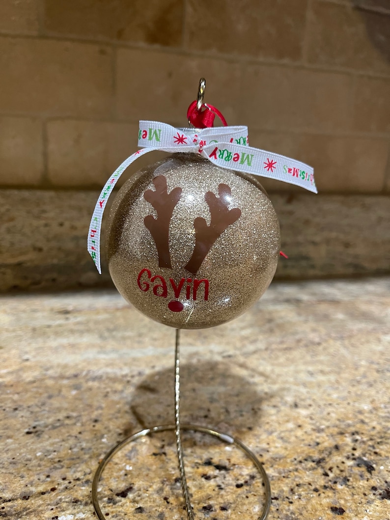 May include: Gold glitter Christmas ornament with a reindeer silhouette and the name "Gavin" written in red. The ornament is on a metal stand with a white ribbon with the words "Merry Christmas" printed on it.