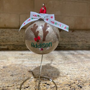 May include: Gold glitter Christmas ornament with a reindeer face and the name Addison. The ornament is on a gold metal stand.