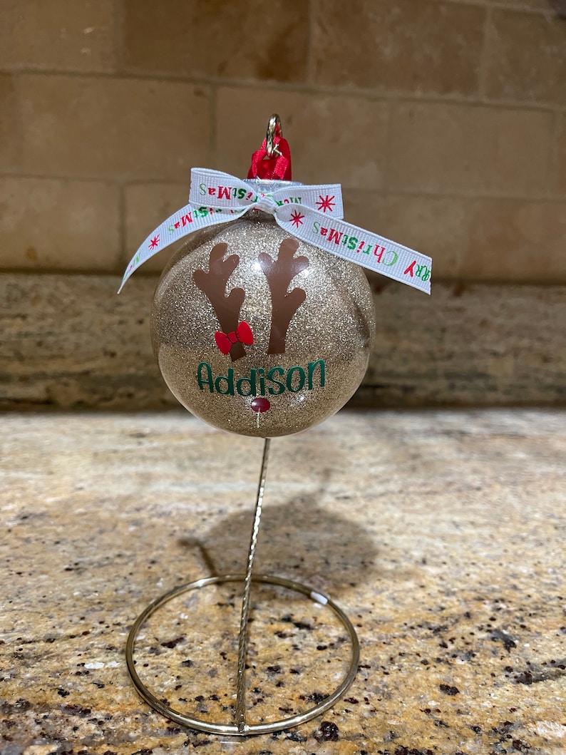 May include: Gold glitter Christmas ornament with a reindeer face and the name Addison. The ornament is on a gold metal stand.