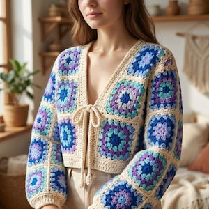 May include: A long-sleeved crochet cardigan featuring a granny square pattern in shades of blue, purple, and teal, bordered by cream-colored yarn. The garment has a deep V-neckline and a front tie closure made of matching cream-colored crochet cord.