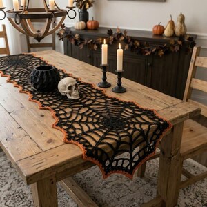 Puede incluir: Un camino de mesa de ganchillo negro con un patrón de telaraña y un borde festoneado de color naranja. El camino está extendido sobre una mesa de madera y decorado con un cuenco negro texturizado y una réplica realista de un cráneo humano.