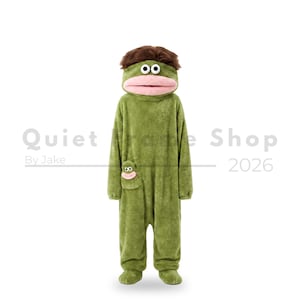 May include: A full-body green plush onesie featuring a character head hood with brown hair, large white eyes, and a wide pink mouth. A small matching character pocket is on the front. The text Quiet Frame Shop By Jake 2026 is visible.
