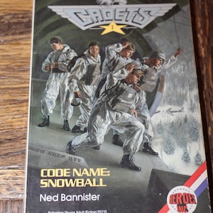 May include: A paperback book titled "Cadets 1" with a cover illustration of young recruits in white uniforms. The book's title is "Code Name: Snowball" by Ned Bannister. The cover features a red, white, and blue logo.